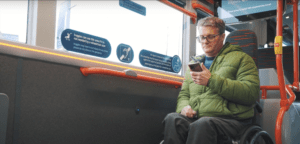 TWC video screenshot bus travel. Wheelchair user using his phone whilst sat on bus
