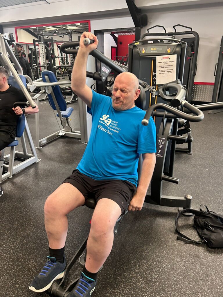 colin phipps in the gym using an arm press whilst wearing an SIA tshirt